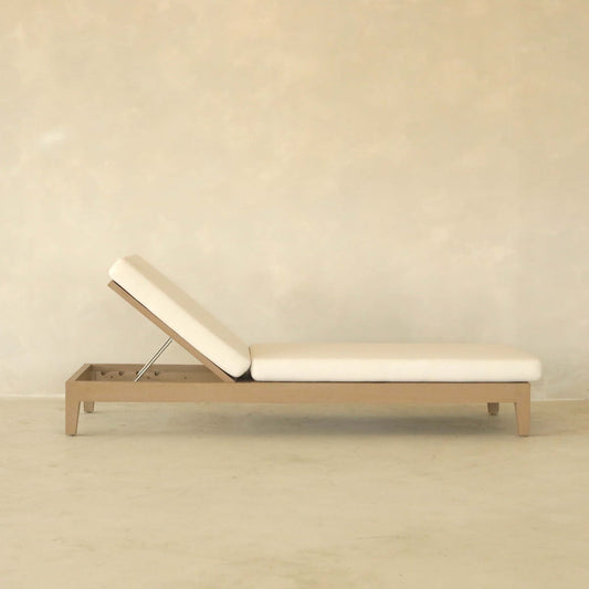 The Bourdain Patio Chaise Lounge features a minimalist beige design with a simple wooden frame and adjustable backrest, providing outdoor comfort. Its shown against a plain, light-colored wall and floor.
