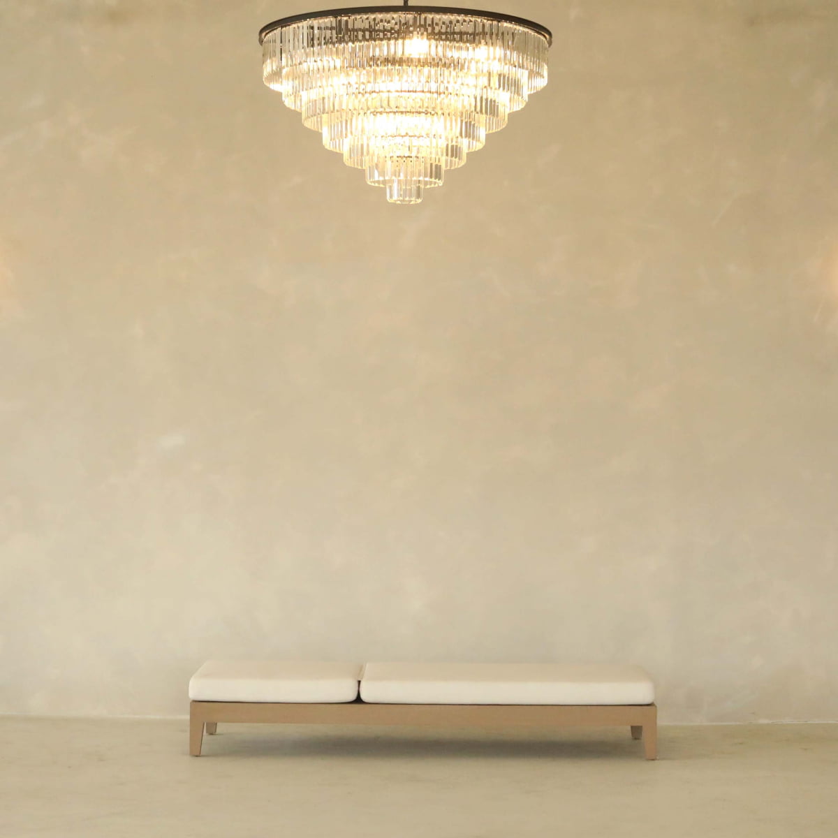 A modern minimalist room showcases a large crystal chandelier and the Bourdain Patio Chaise Lounge, a sleek, light-colored bench with ergonomic design, positioned against a beige wall.