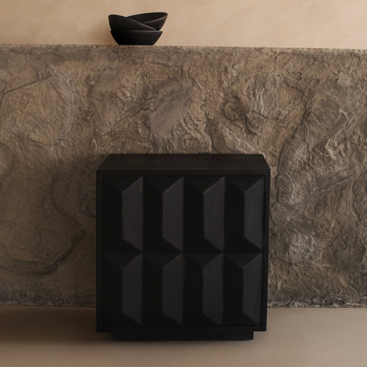 The sleek black Braemar Console with a geometric design stands before a textured stone wall, while two stacked black bowls on the stone ledge above complete the minimalist, contemporary look.