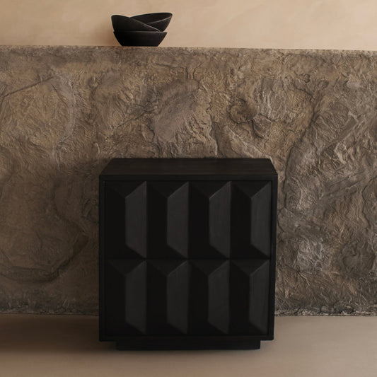 The sleek black Braemar Console with a geometric design stands before a textured stone wall, while two stacked black bowls on the stone ledge above complete the minimalist, contemporary look.