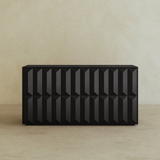 The Braemar Console Large, designed by Anthony Newman, features a modern black finish with a geometric textured front, standing out against a simple beige wall and floor for an avant-garde look.