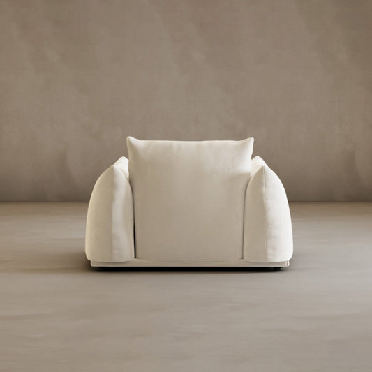The Calypso Arm Chair boasts plush comfort and modern elegance with wide, rounded arms and a thick back cushion. Set on a smooth beige floor, it stands out against a plain light taupe wall.