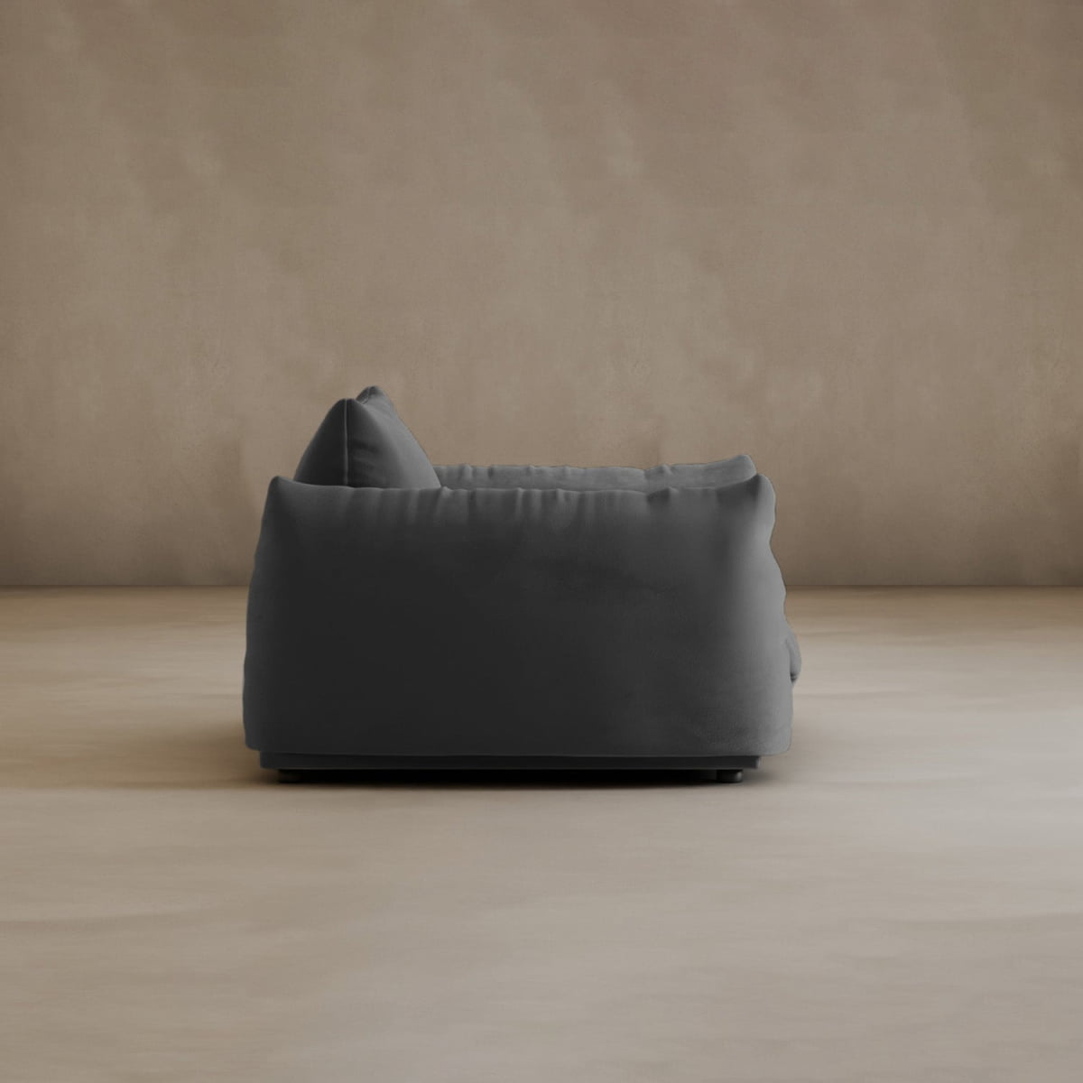 A side view of the Calypso Arm Chair highlights its modern elegance, with plush comfort and soft, rounded edges in dark gray set against a plain beige wall and floor.