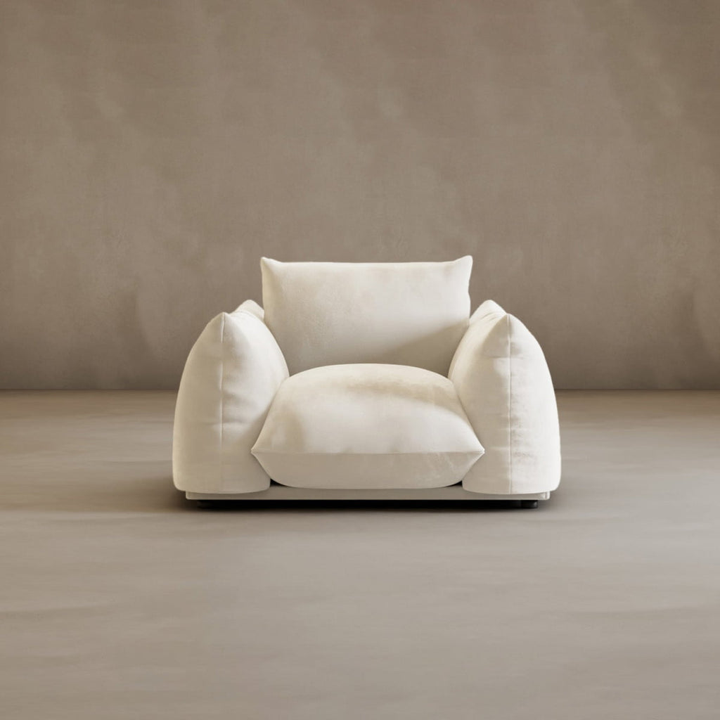 The Calypso Arm Chair, featuring soft, rounded cushions and plush white upholstery, sits against a taupe background. Its oversized design and forward-facing position showcase its modern elegance and inviting comfort.