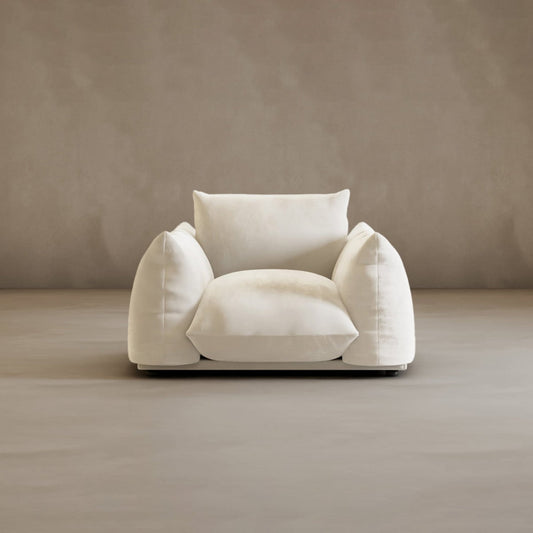 The Calypso Arm Chair, featuring soft, rounded cushions and plush white upholstery, sits against a taupe background. Its oversized design and forward-facing position showcase its modern elegance and inviting comfort.