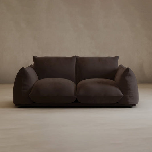 The Calypso Loveseat, a modern 2-seater in dark brown with overstuffed seat and back cushions, stands out against a beige wall and light floor, combining contemporary style with plush comfort.