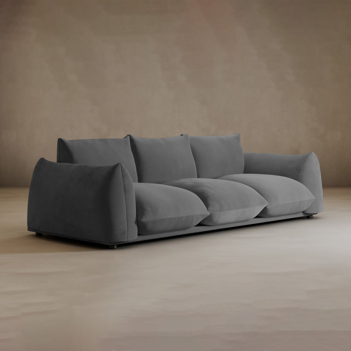 A modern Calypso Three Seater Sofa in deep gray, featuring plush, oversized cushions, sits in a minimalist room with neutral beige walls and floors—ideal for contemporary living spaces.