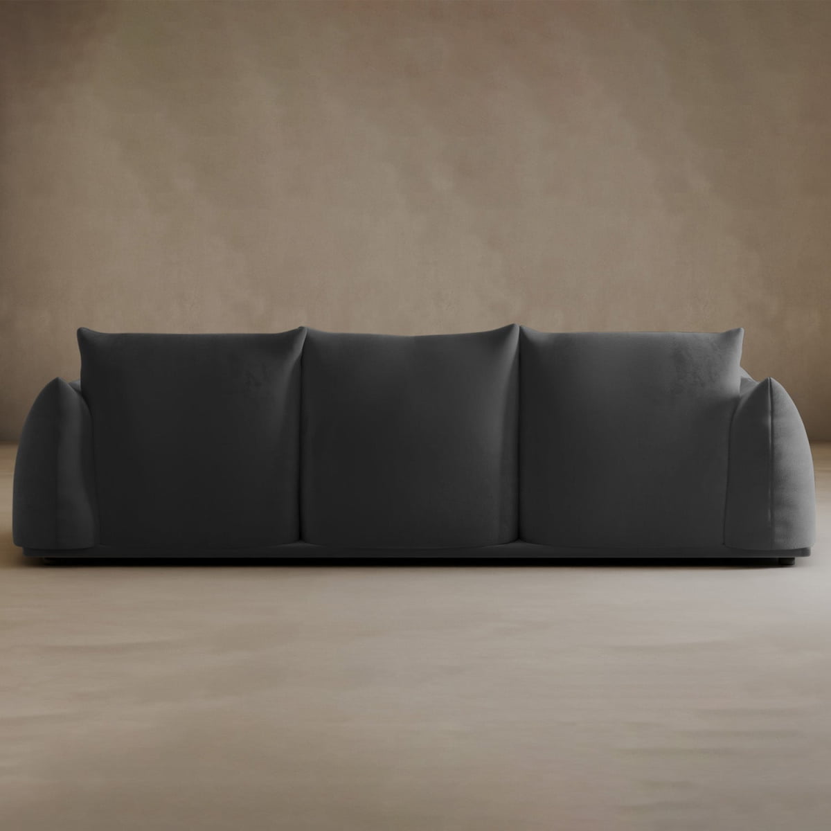 The Calypso Three Seater Sofa, a modern minimalist piece in dark gray with three wide cushions and rounded armrests, stands against a plain beige wall and floor.