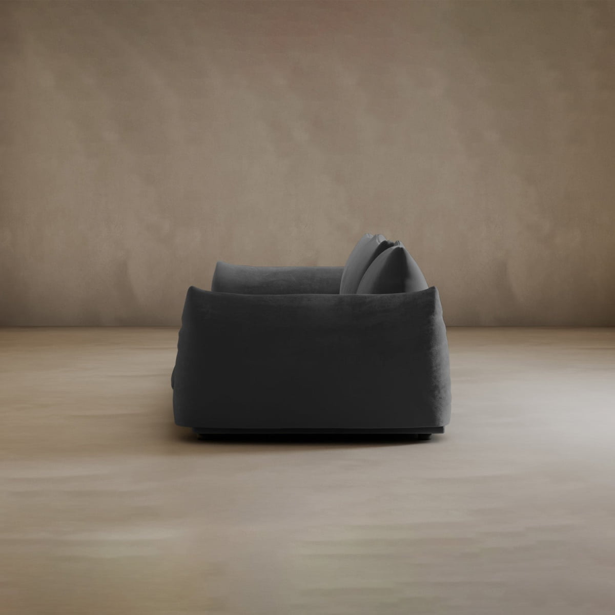 The Calypso Three Seater Sofa in dark gray with plush cushions is shown side-on in a minimal room with beige walls and a light floor—a chic, modern choice for your living space.