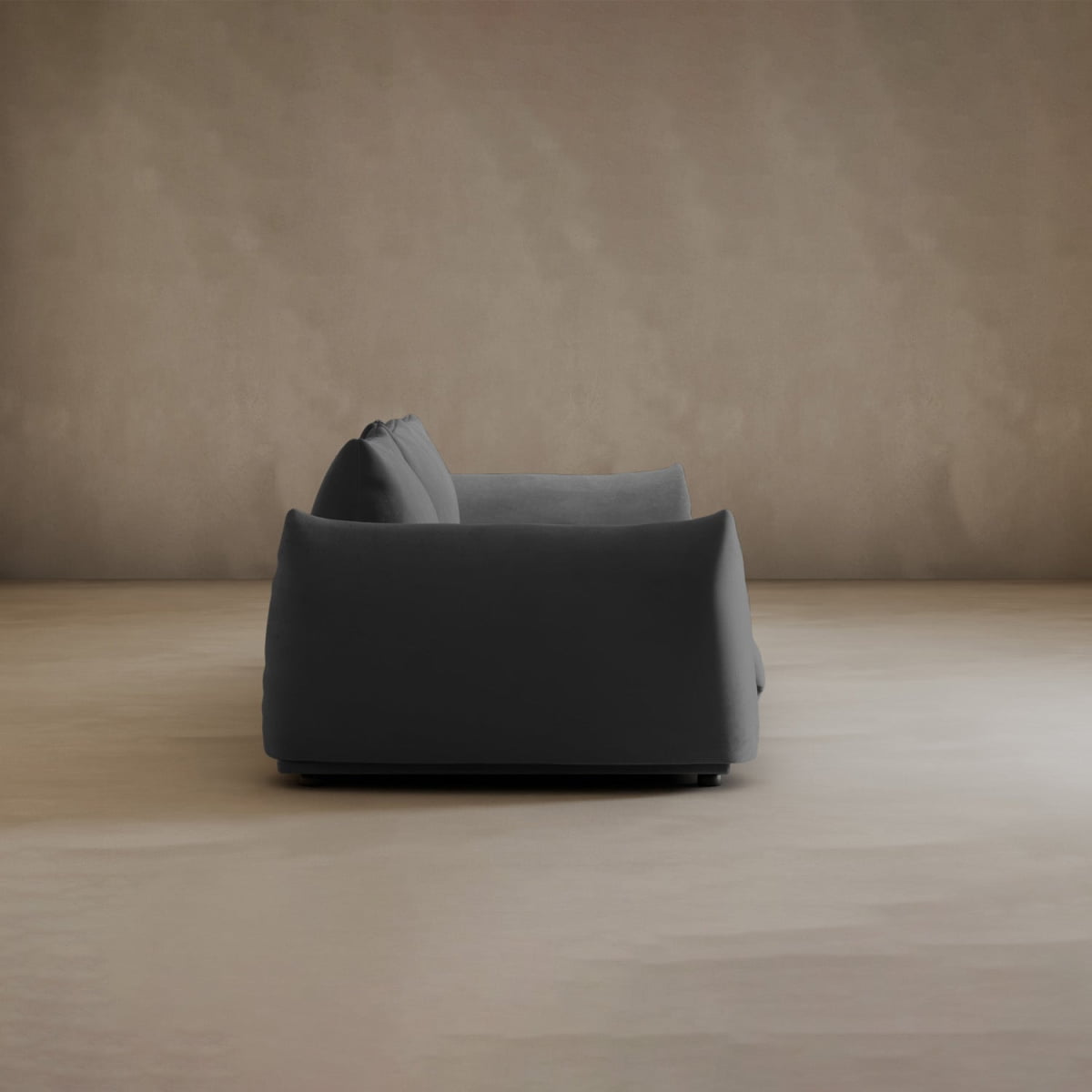 A modern Calypso Three Seater Sofa in dark gray is seen from the side, placed against a plain beige wall and floor in a minimalist, empty room.