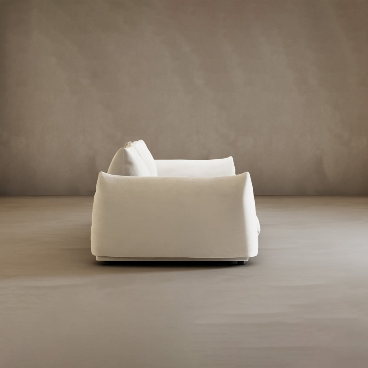 The Calypso Three Seater Sofa features plush comfort and modern elegance, shown side-on in a minimalist room with a smooth beige wall and matching floor, its white upholstery enhancing the serene setting.
