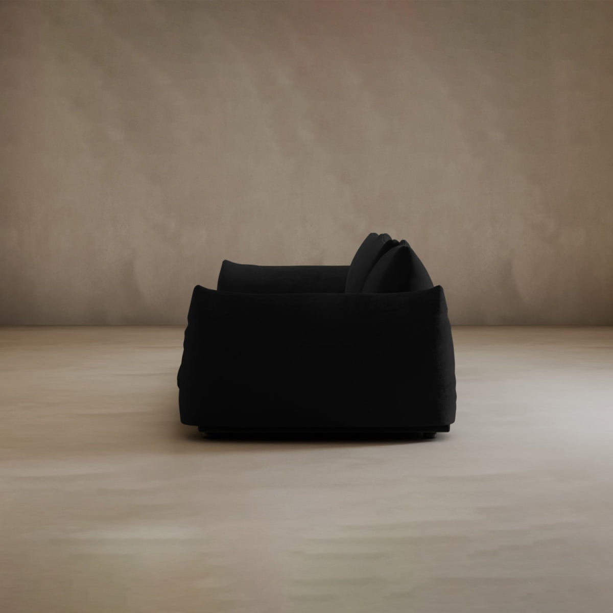 A black Calypso Three Seater Sofa sits centered in a minimal room with beige walls and a light floor, its sleek design highlighted by the uncluttered space.