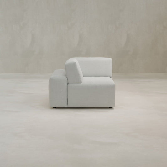The Canonbury Corner, a minimalist light gray upholstered sofa chair, sits on a light floor against a textured beige wall, bringing contemporary comfort to any space.