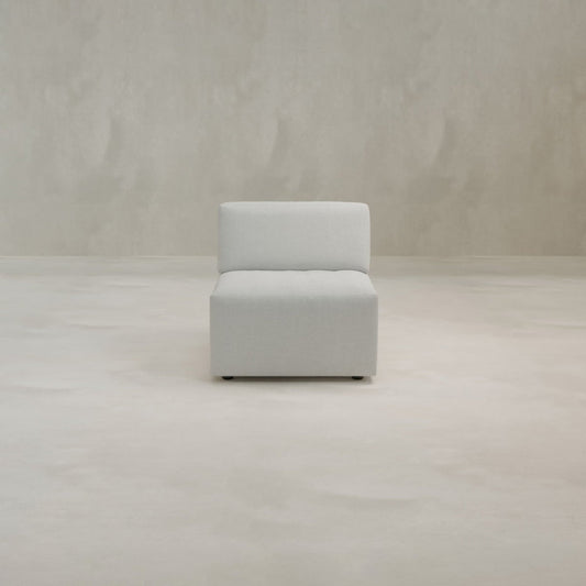 The Canonbury Middle Seat, an armless chair in light gray upholstery, is centered in a bare room with a matching gray floor and textured wall, highlighting its contemporary comfort and timeless style.