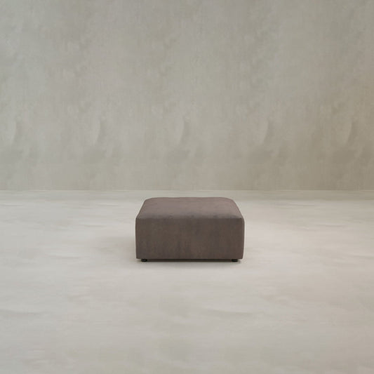 The Canonbury Ottoman, a square, dark brown piece in high-performance fabric, is centered on a light floor against a neutral wall, highlighting its modern style and contemporary comfort.