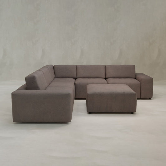 The Canonbury Sofa Set With Ottoman features a large, modern L-shaped brown upholstered sectional with wide seats and low backs, plus a matching square ottoman, all set against a plain light gray wall and floor for contemporary comfort.
