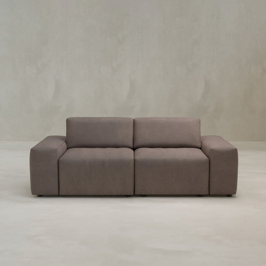 The Canonbury Two Seater is a modern, minimalist brown sofa with high-performance fabric, wide armrests, and rectangular cushions, placed on a light floor against a plain light gray wall.