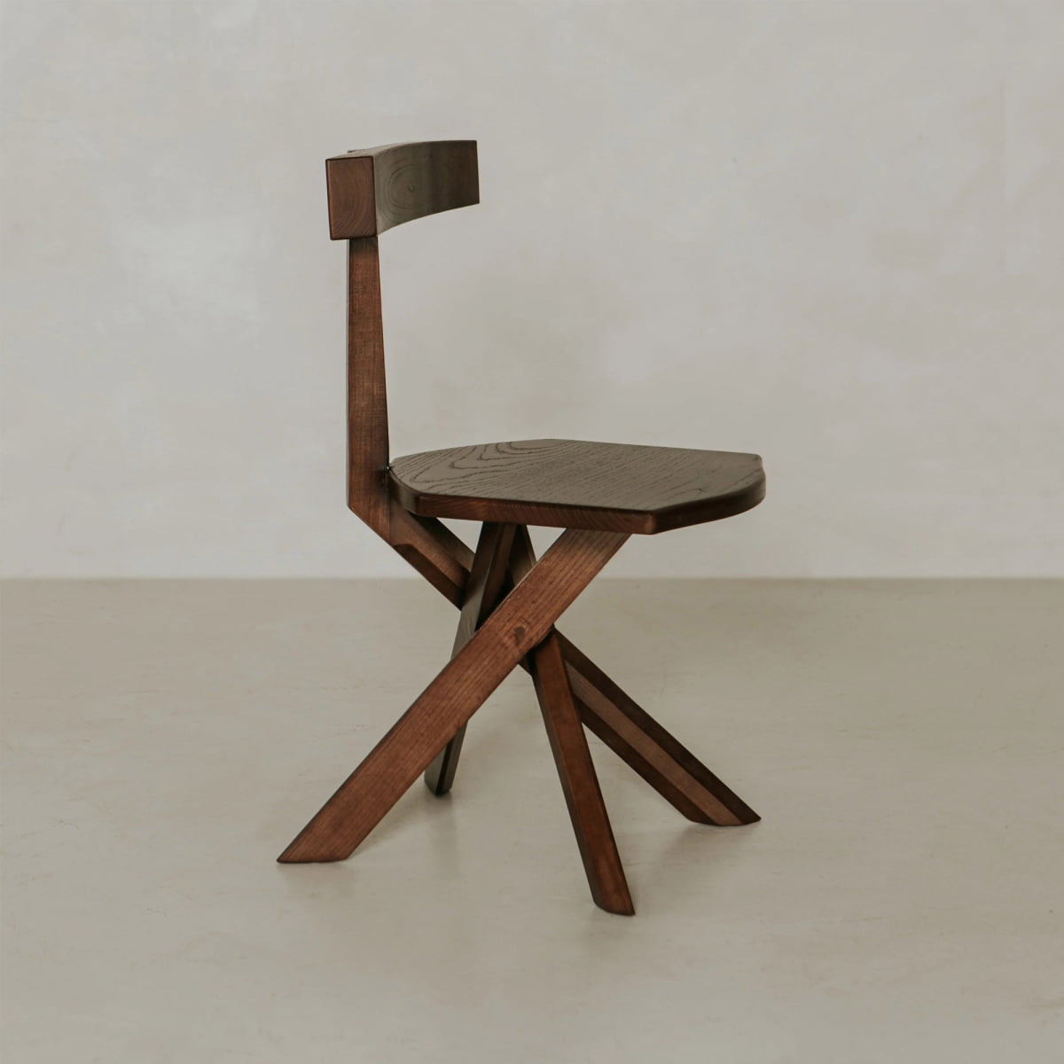 The Cartel Chair is a modern wooden chair with bold angles, a flat seat, narrow backrest, and crossed legs in an angular geometric style, shown against a plain off-white background.