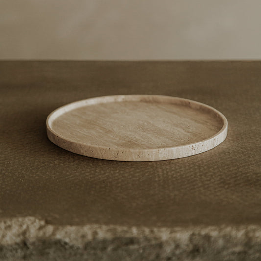 The Cascade Tray in Small, a round flat travertine piece, rests on a textured brown surface with a neutral backdrop, adding natural elegance to any decorative display.