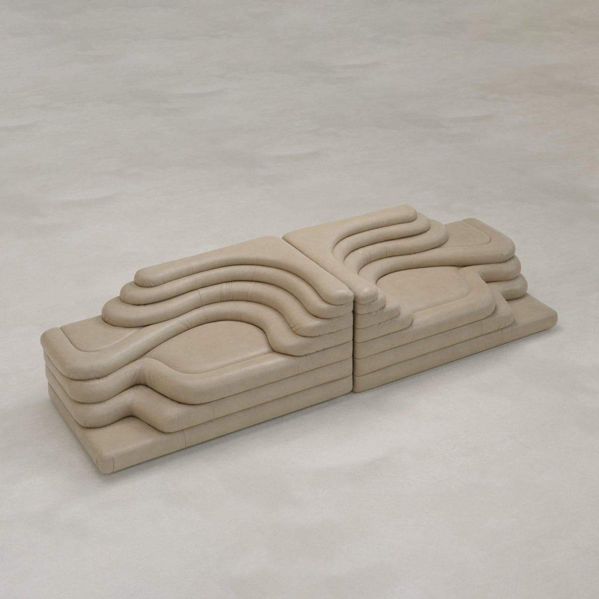 The Cascata 2 Piece Sofa Rental features sculptural, beige seating with layered, wavy contours forming two mirrored sections, creating a striking modern statement on a smooth light floor.