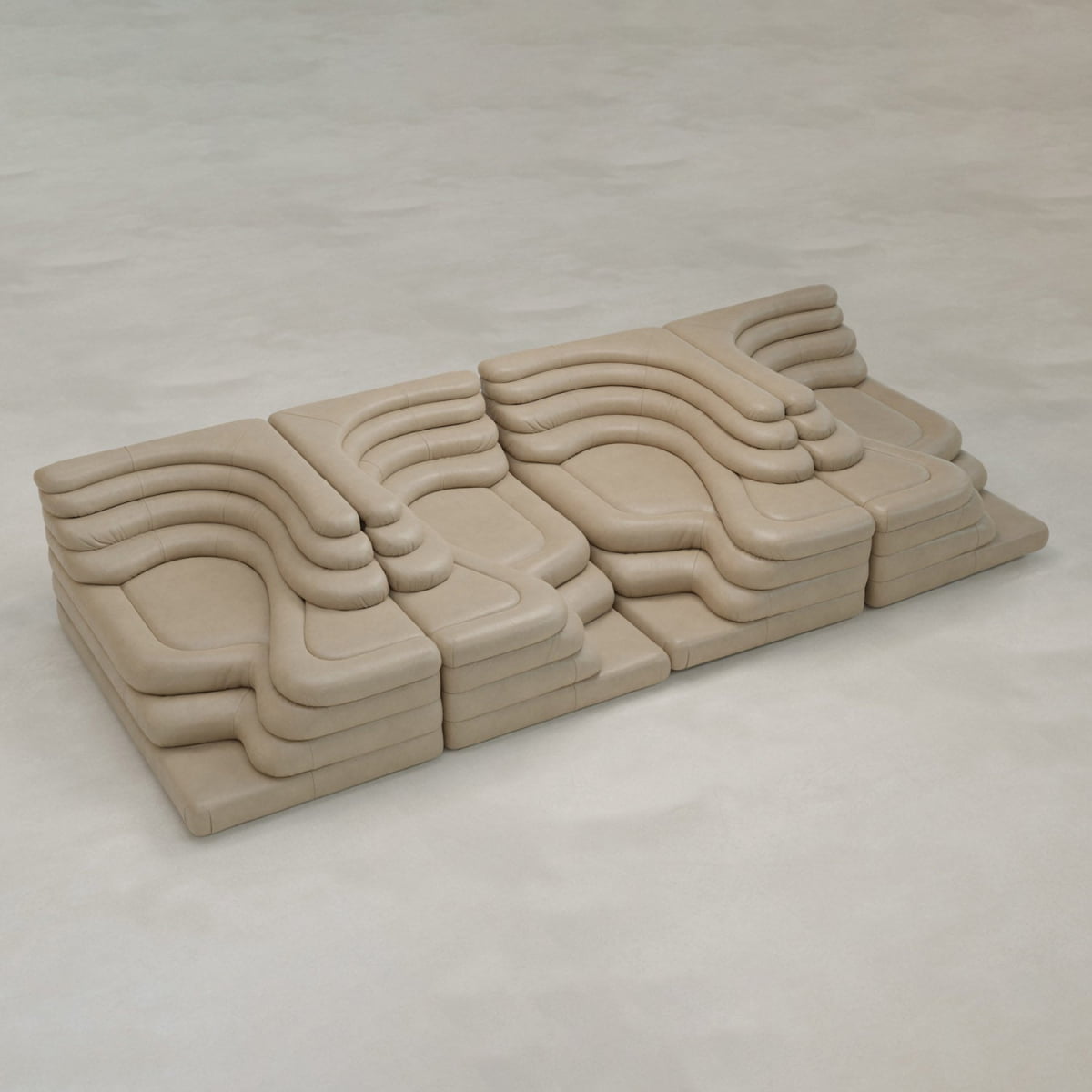 The Cascata 4 Piece Sofa Rental features modular beige sections with layered, sculptural cushions in wavy shapes, offering a luxurious look on a light floor in a minimalist space.