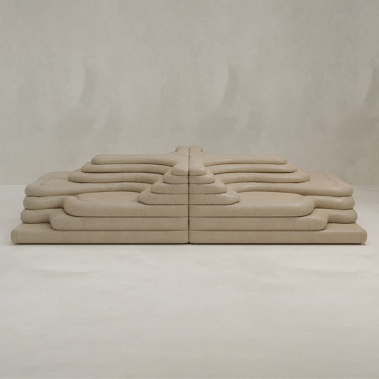 The Cascata 4 Piece Sofa Rental features a modular, sculptural design with layered, rounded beige cushions arranged symmetrically against a neutral wall and floor, perfectly embodying modern style.