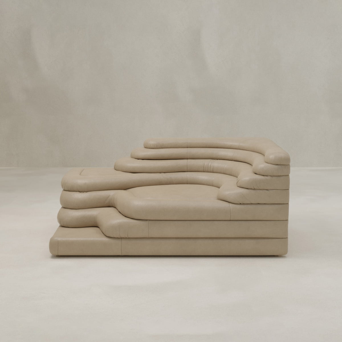 The Cascata Sofa Left Arm is a sculptural beige leather modern sofa featuring layered, wave-like cushions and a sleek design, perfectly complementing a neutral room with plain light walls and flooring.