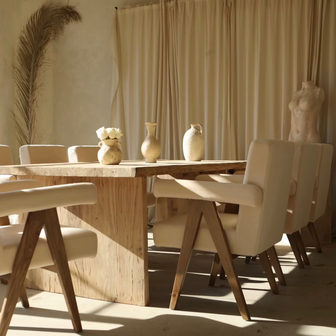 A sunlit dining room features a wooden table, six Chelsea Dining Chairs with vegan upholstery, ceramic vases, a dried palm leaf, beige curtains, and a torso sculpture for a minimalist, natural look.