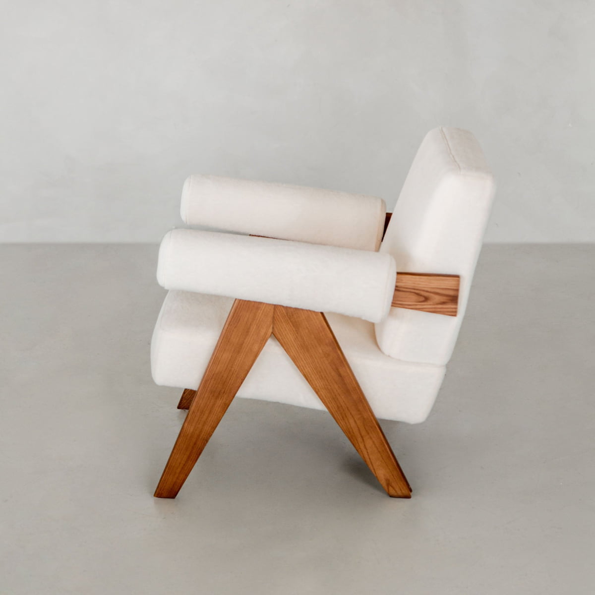 The Chelsea Lounge Chair features white faux mohair upholstery and wooden legs, presenting a refined mid-century look in a minimalist space with a light gray wall and floor.