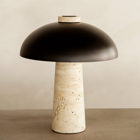 The Chiara Lamp features a textured, cylindrical beige travertine base paired with a smooth, matte black dome shade, emphasizing its modern design against a neutral backdrop.