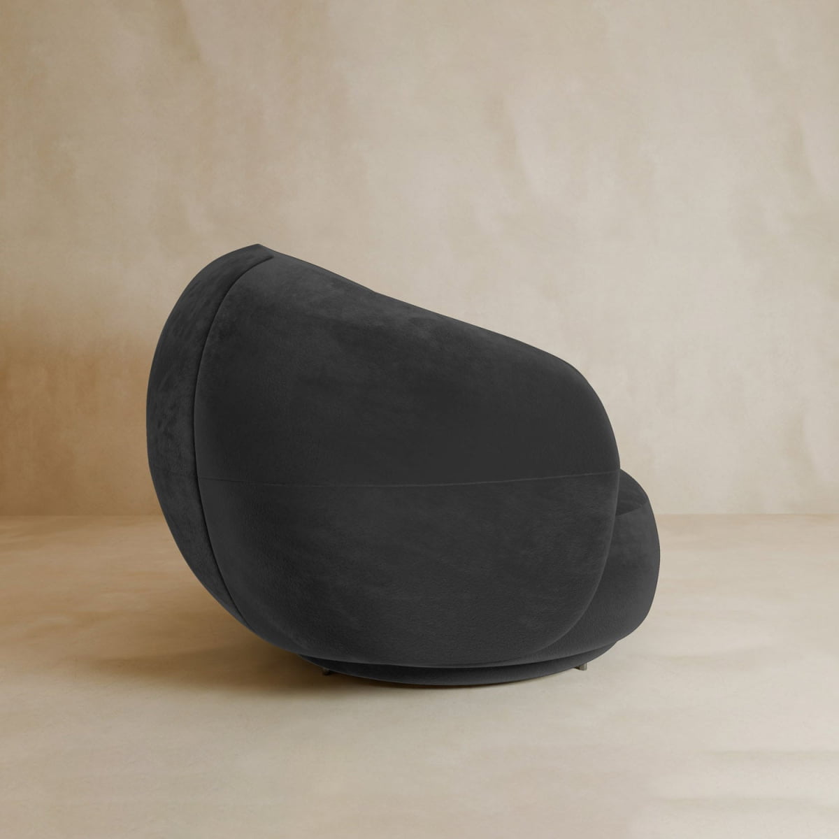 The Clara Armchair features plush black velvet upholstery, a rounded cocoon-like shape, and a soft, smooth texture, adding contemporary style to any space. It sits on a light beige floor against a matching beige wall.