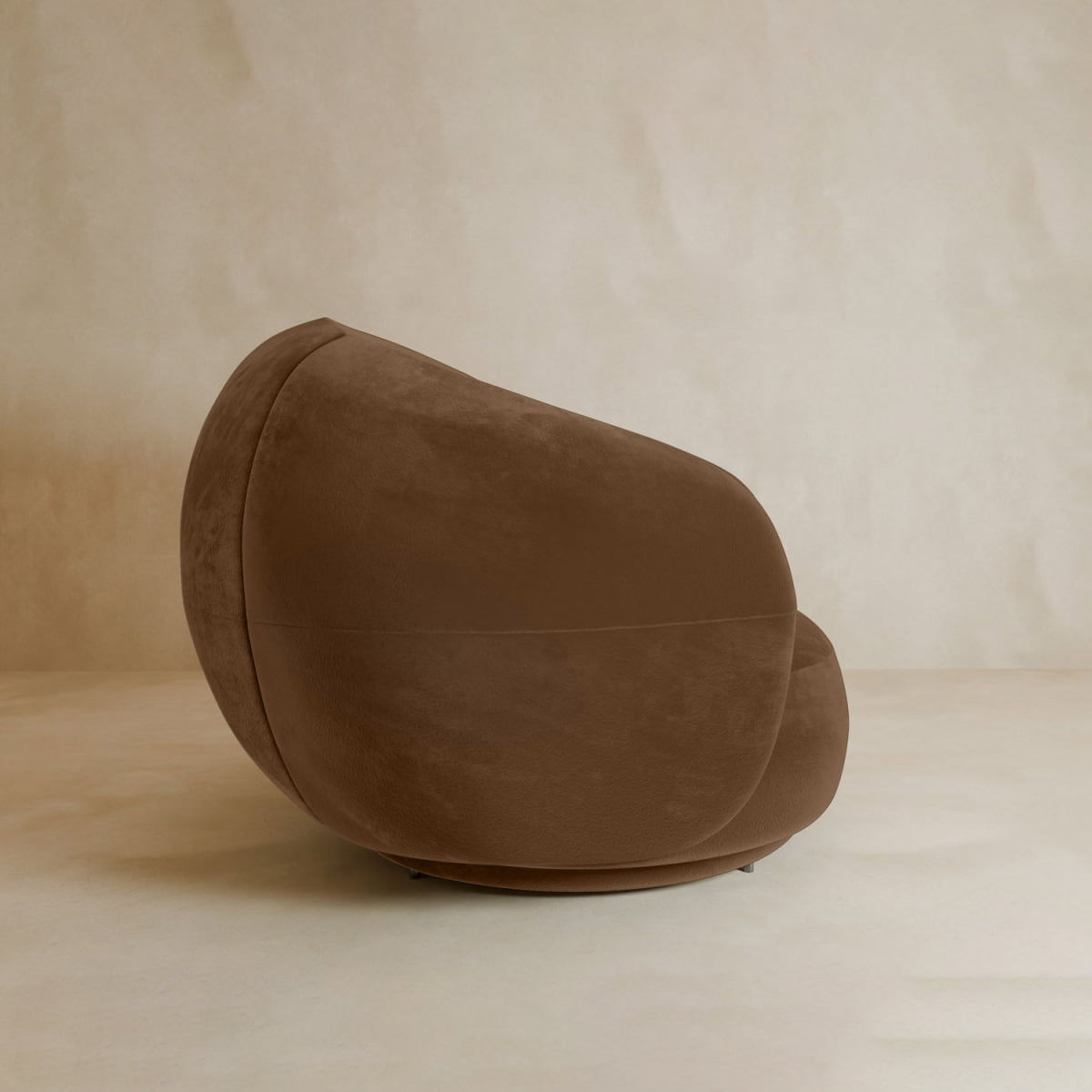 The Clara Armchair features plush brown velvet upholstery and rounded contours, set on a light beige floor with a matching wall, offering contemporary style and minimalist comfort.