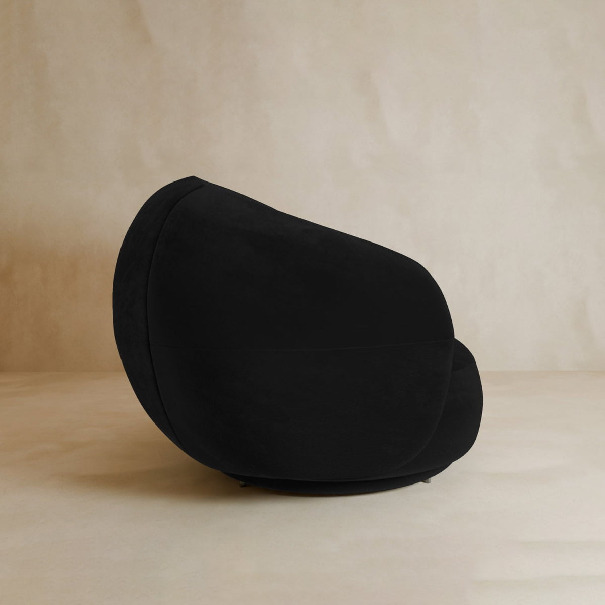 The Clara Armchair, featuring a modern black design with velvet upholstery and smooth sculptural lines, is placed on a beige floor against a pale textured wall.