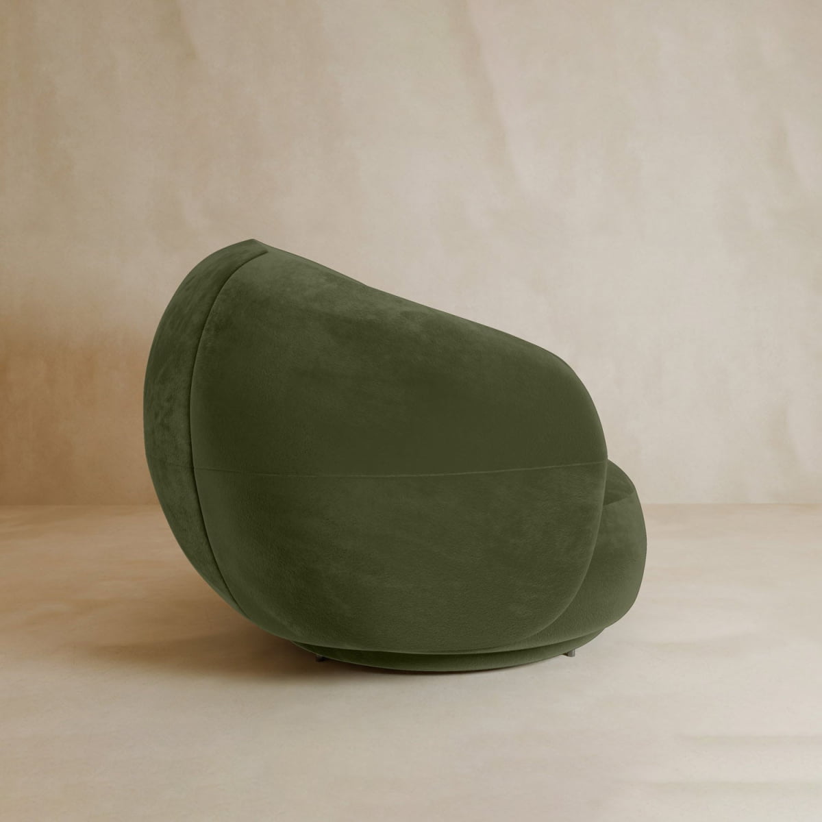 The Clara Armchair features a modern, sculptural design with luxurious green velvet upholstery and rounded contours, positioned on a beige floor against a matching beige wall.