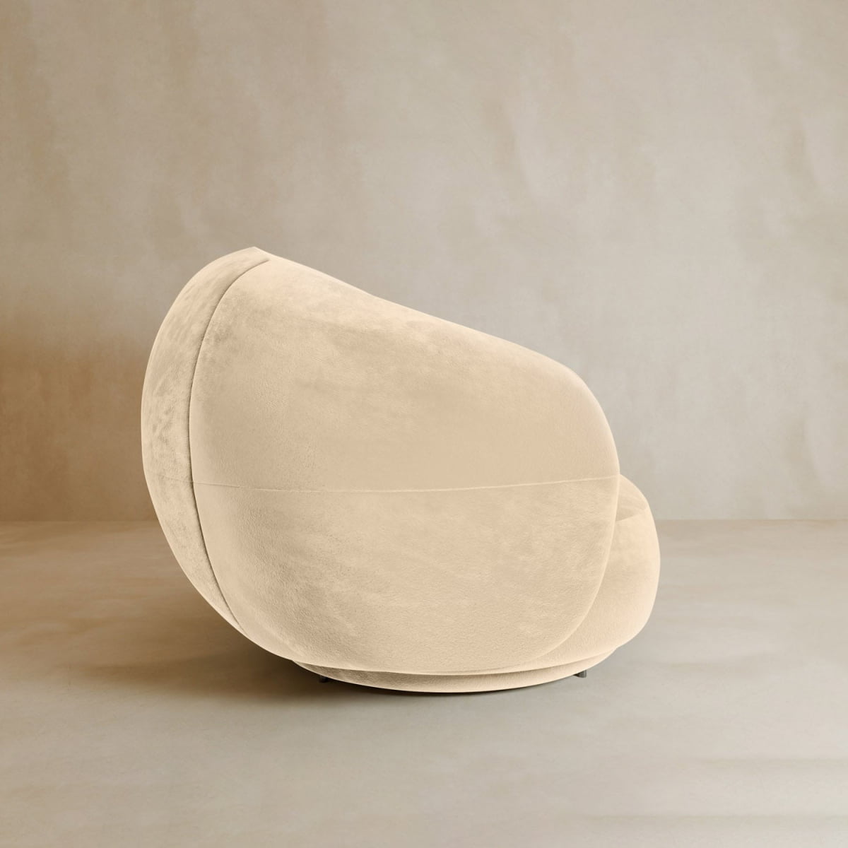 The Clara Armchair features plush velvet upholstery and a rounded, sculptural design, positioned on a smooth neutral floor against a light-colored wall.