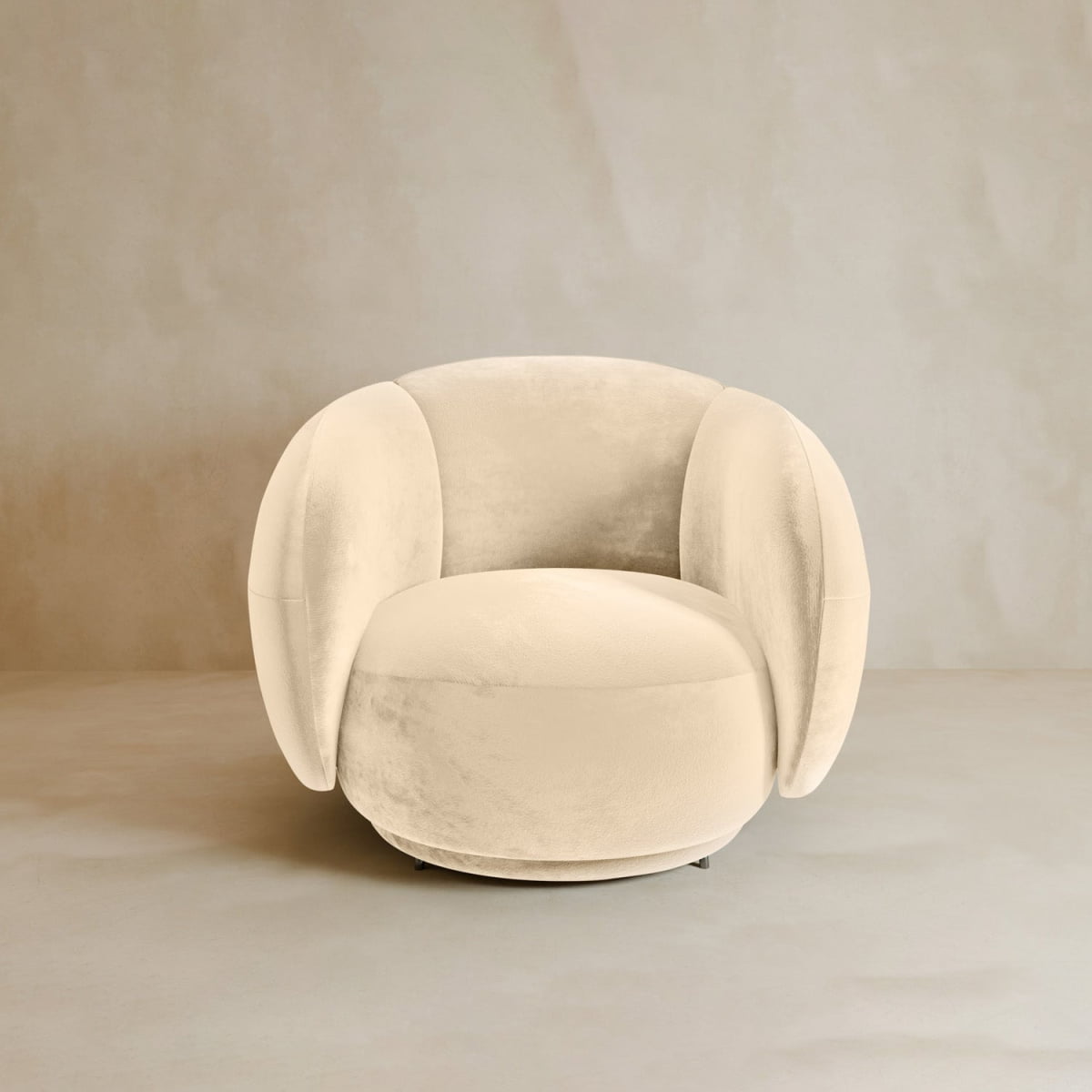 The Clara Armchair, a plush round beige seat with curved armrests and backrest in contemporary style, features soft velvet upholstery and sits on a neutral floor against a light beige wall.