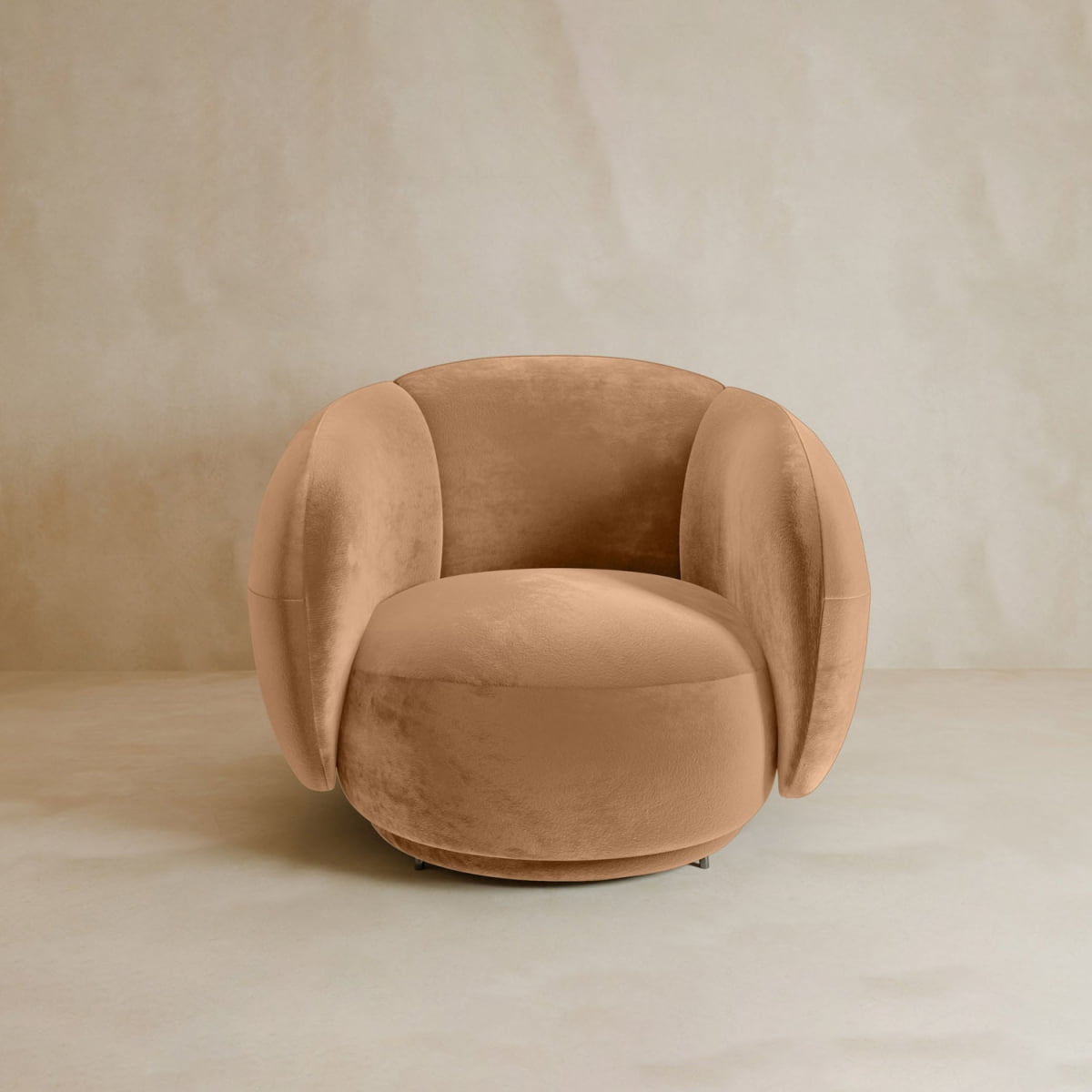 The Clara Armchair, with its plush contemporary design, features soft tan velvet upholstery, rounded armrests, and a curved backrest for a cozy look. It sits on a neutral textured surface against a light beige wall.