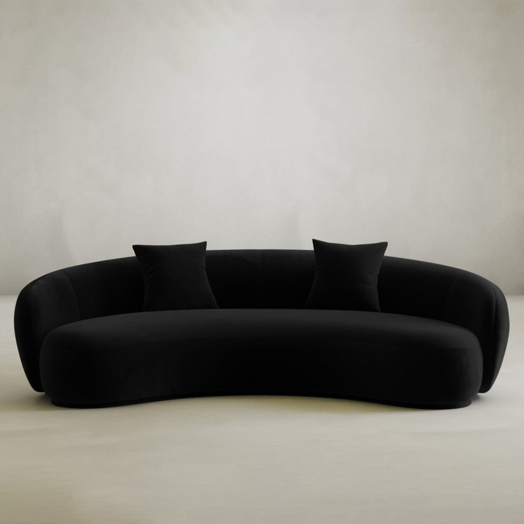 The Clara Sofa, a modern curved black piece with two matching throw pillows, adds style and comfort to any contemporary home against a neutral backdrop.