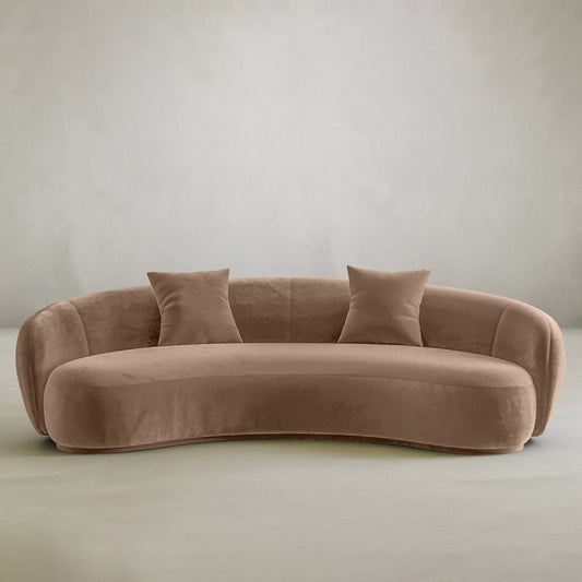 The Clara Sofa features a modern curved design in light brown velvet with two matching cushions, perfect for a contemporary home. Shown against a plain, light grey background.