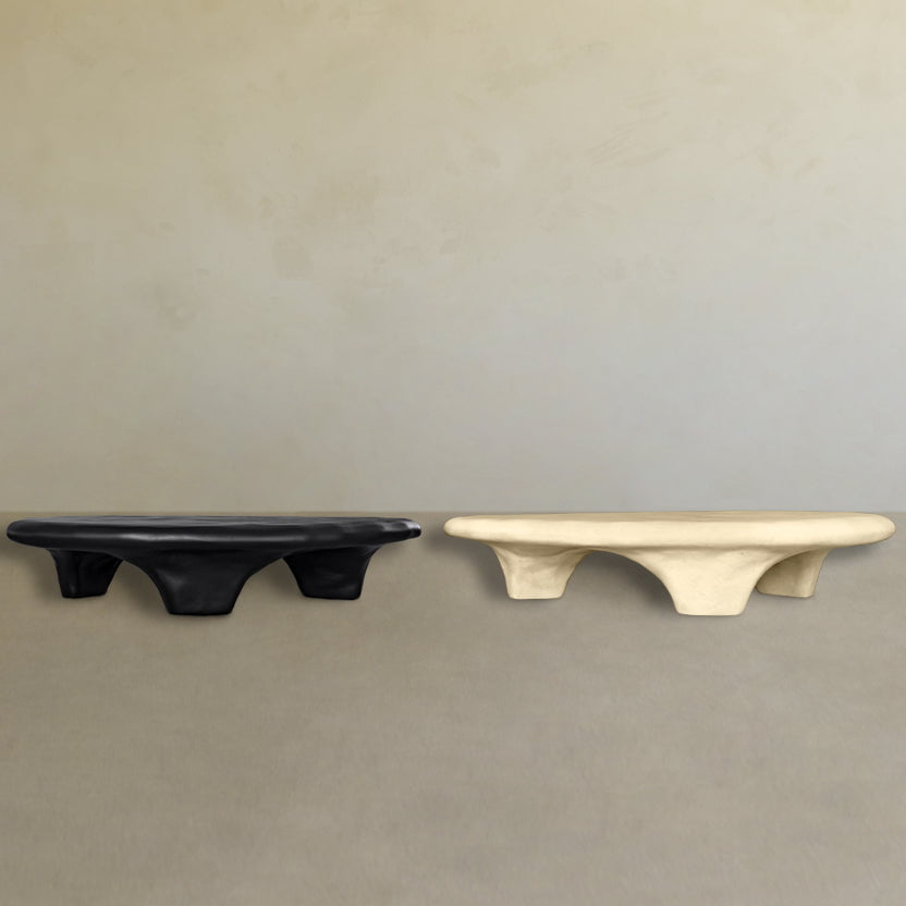 The Cleo Coffee Table features a low, sculptural design with four thick legs. Shown in matte black and off-white finishes, both tables have rounded edges and are set on a neutral floor against a light beige wall.