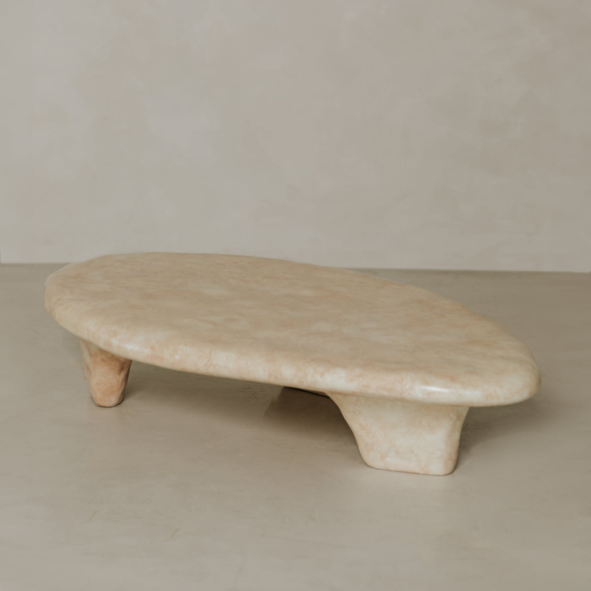 The Cleo Coffee Table features an oval, beige stone design with two short, wide legs, set against a matching beige wall and floor. Its smooth, polished surface and minimalist form subtly reference brutalist style.