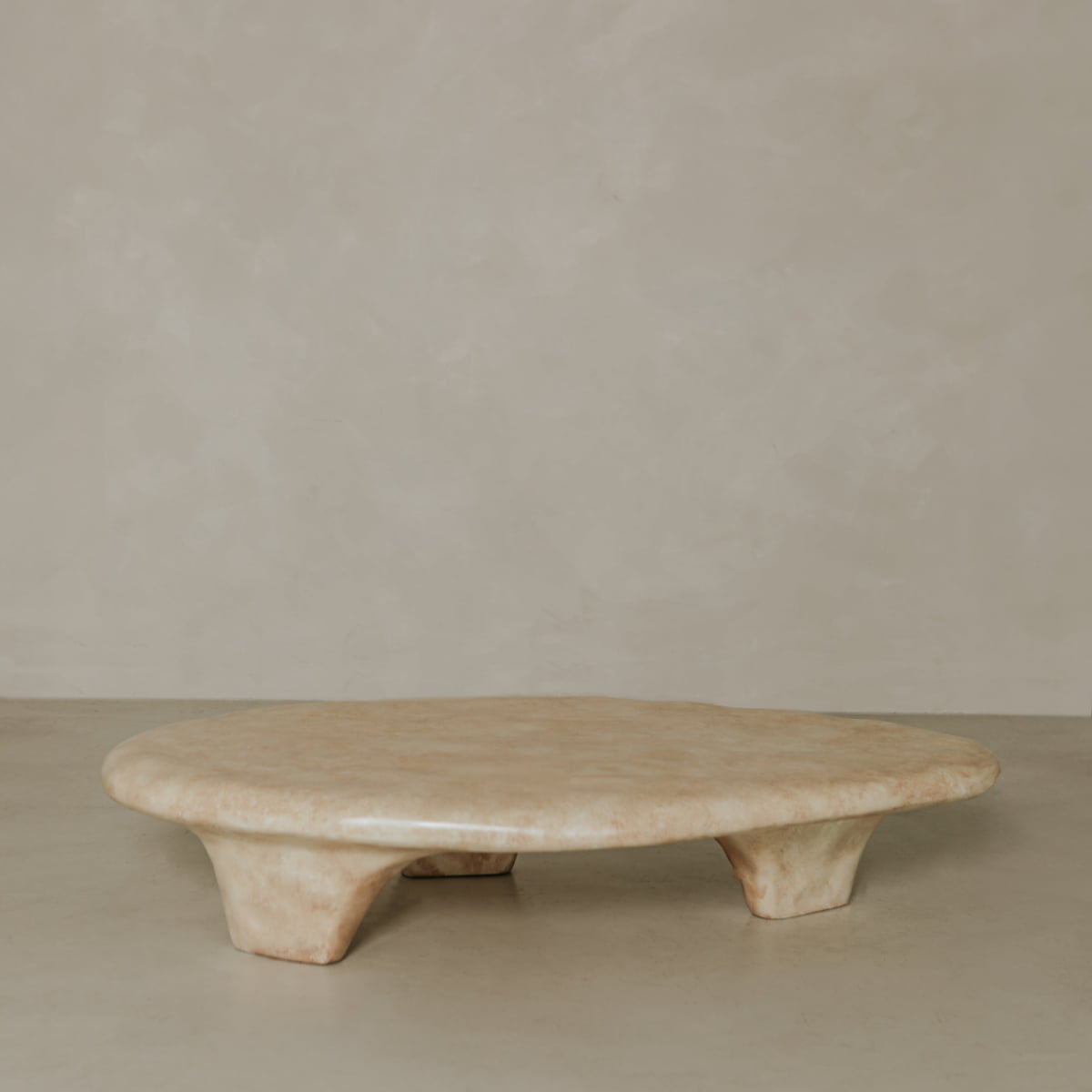 The Cleo Coffee Table is a low, oval-shaped beige piece with three short, wide legs, inspired by natural forms and displayed against a plain light-toned wall and floor.