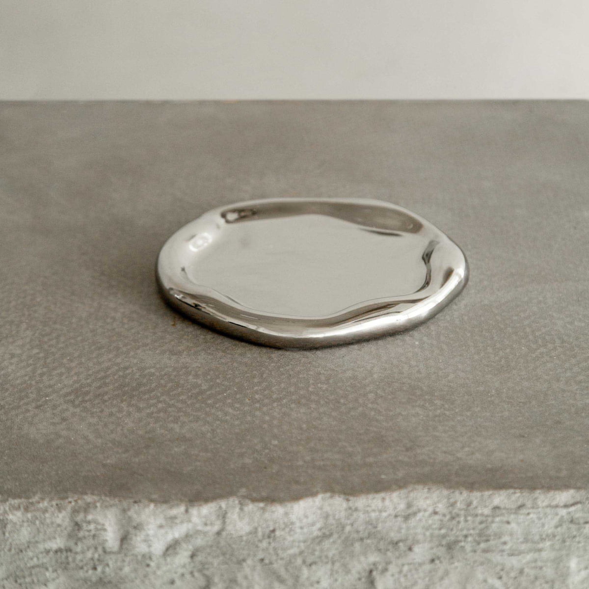 The Coda Tray, a small silver plate with an irregular, wavy edge, rests on a textured gray stone surface, radiating contemporary elegance.