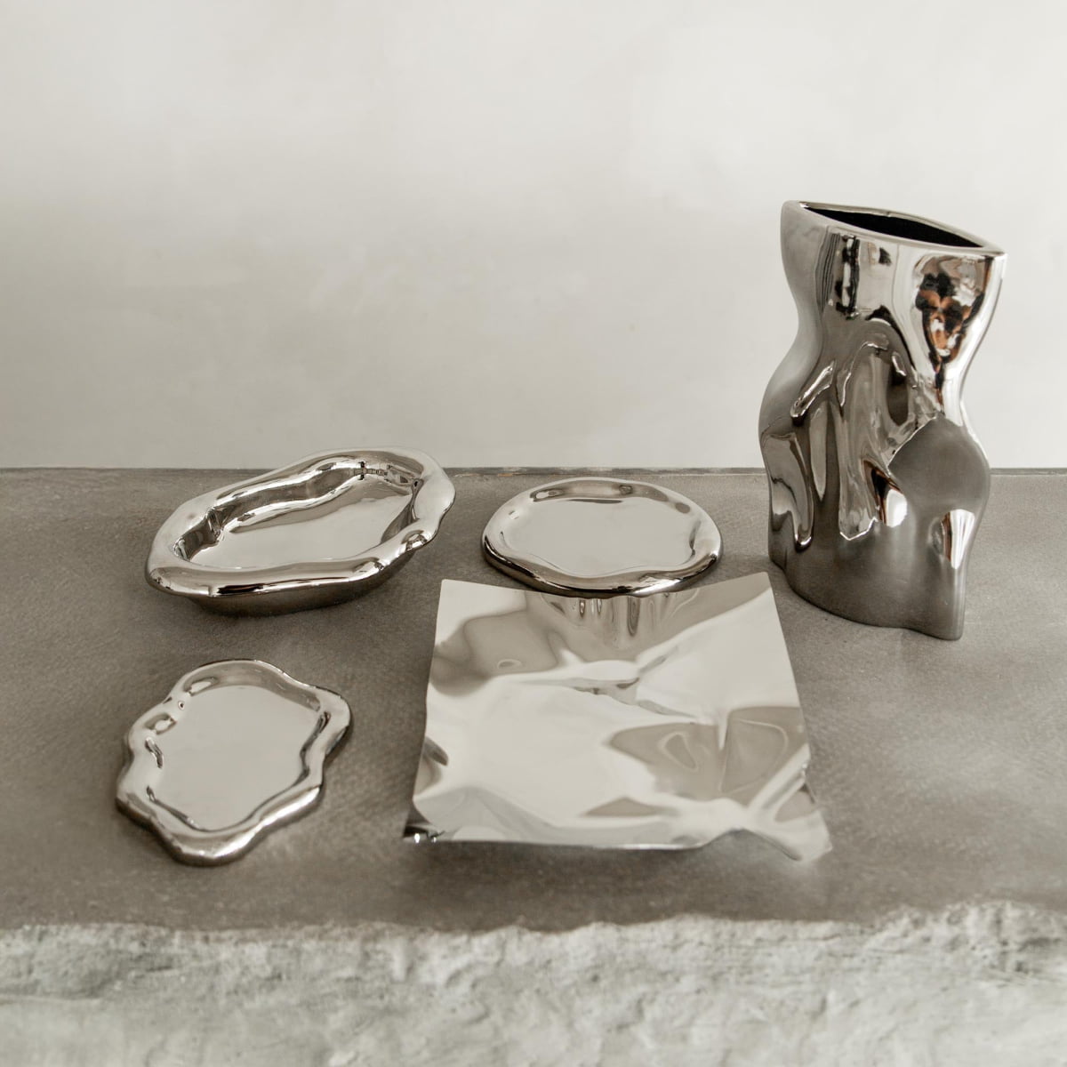Five abstract, silver metallic Coda Trays with organic, uneven shapes are displayed on a gray textured surface against a plain light background, featuring varied forms including plates and a tall, wavy vase.