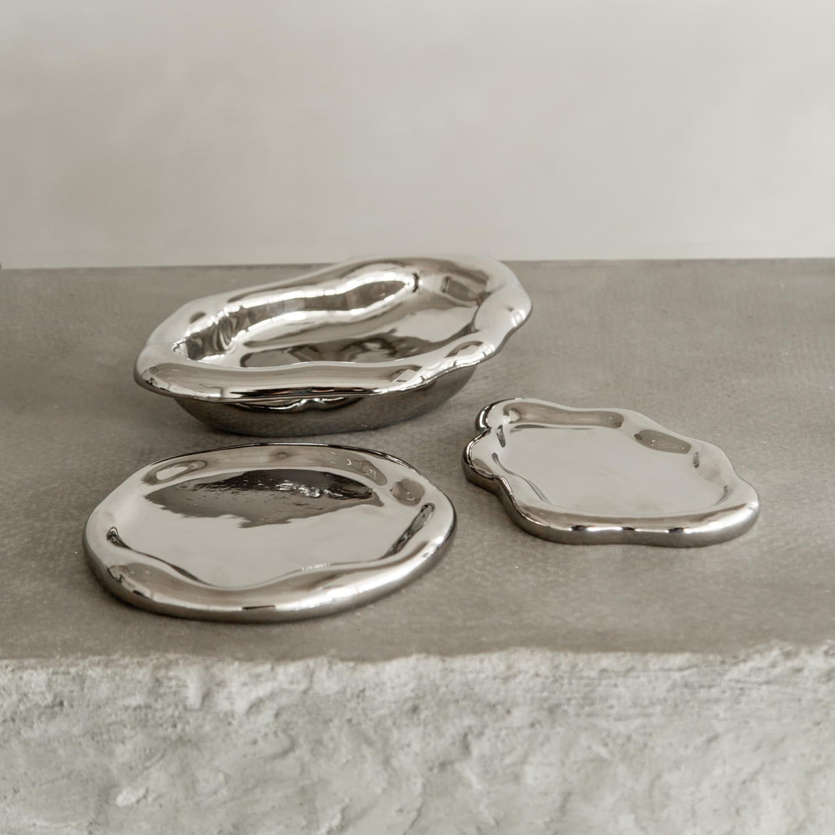 Three Coda Trays, featuring an irregular wavy silver design, are displayed on a textured light gray stone surface with a plain neutral background.