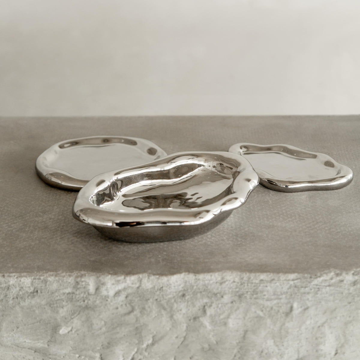 Three Coda Trays, featuring irregular shapes and reflective silver finishes with wavy edges, bring contemporary elegance as they rest on a textured light gray surface against a plain light background.