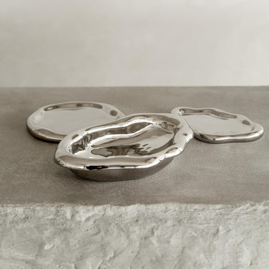 Three Coda Trays, featuring irregular shapes and reflective silver finishes with wavy edges, bring contemporary elegance as they rest on a textured light gray surface against a plain light background.