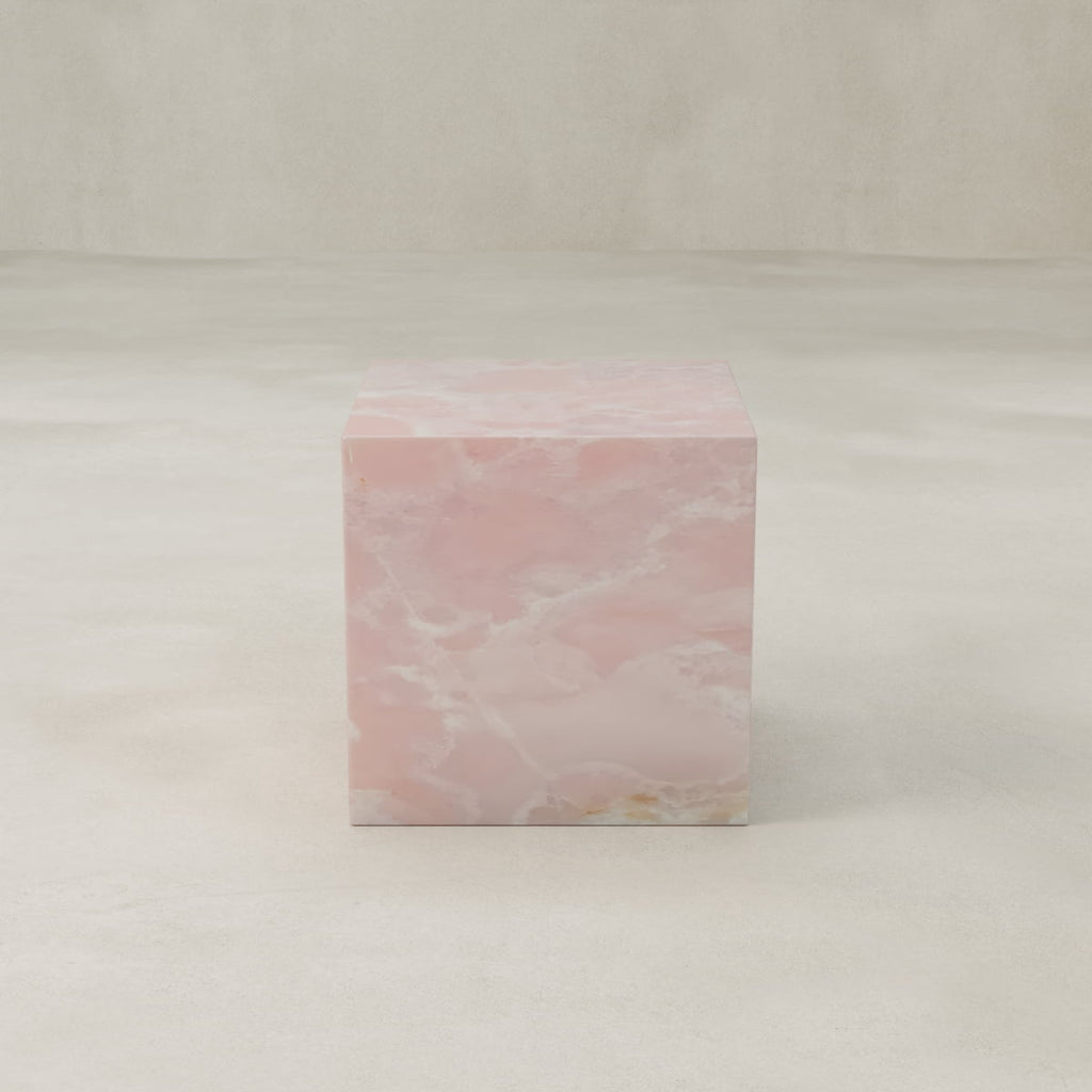 The Cora Plinth, a square pink onyx pedestal with a smooth surface and cloudy natural patterns, rests on a light textured floor against a neutral backdrop.