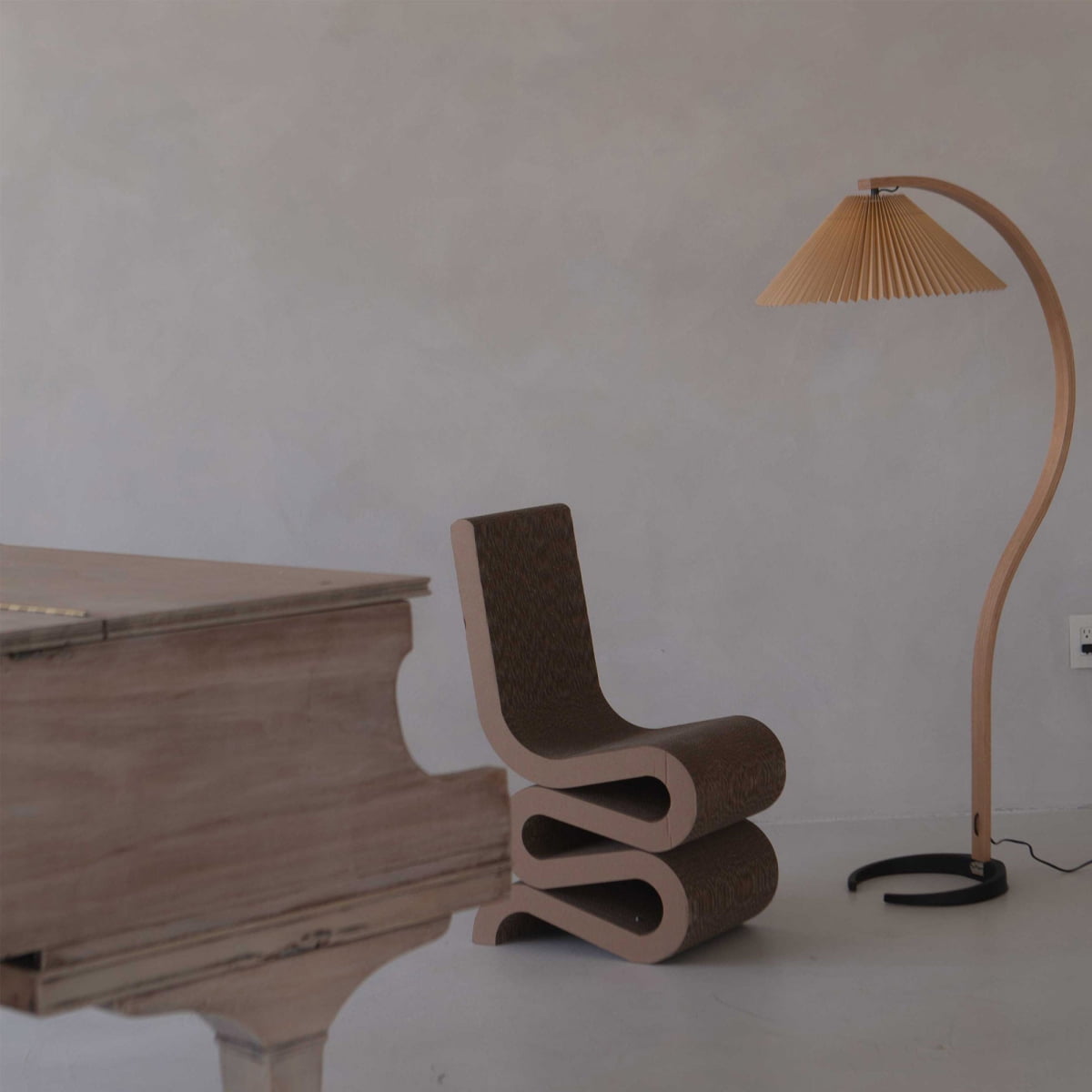 A minimal room featuring contemporary pieces: the Curly Wurly Chair, a curving floor lamp with a pleated shade, and the corner of a wooden piano—set against a plain light-colored wall.