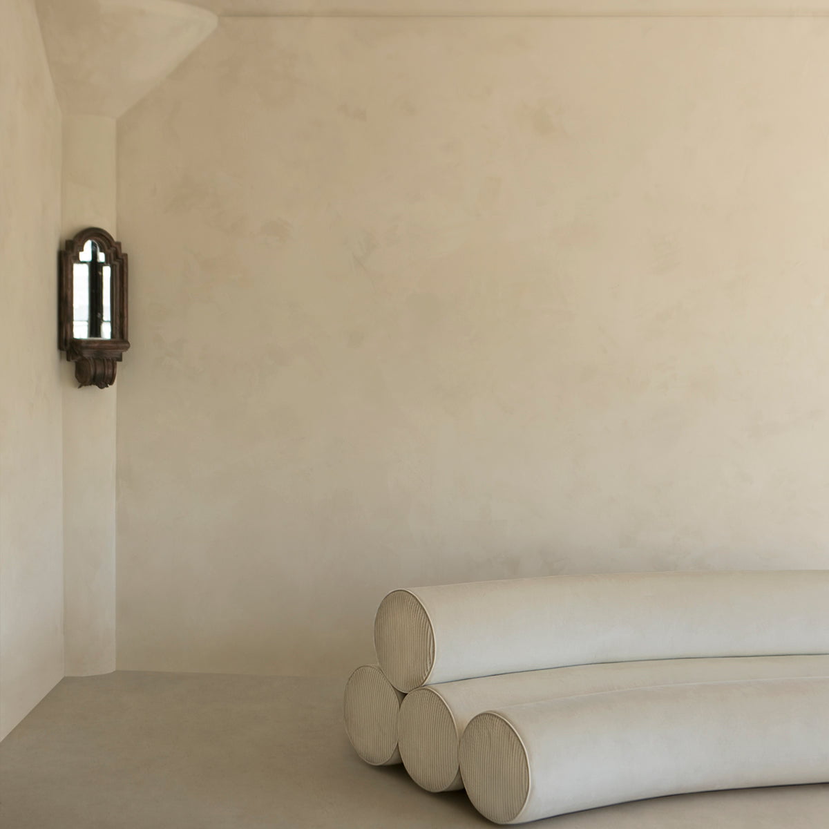 Minimalist room with a textured beige wall, vintage sconce on the left, and four stacked Cylindra Sofa Rentals in the corner on a plain floor—ideal for modern spaces.